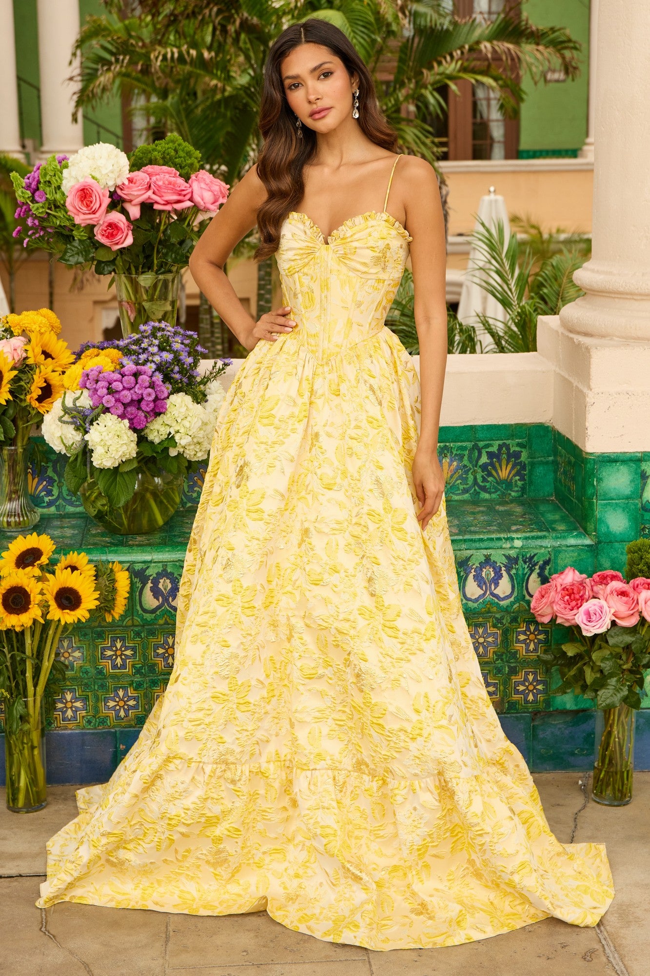 A woman wears the Ava Presley 42312 long yellow prom dress with an elegant A-line skirt, standing outdoors among green tiles, columns, and vibrant flowers.